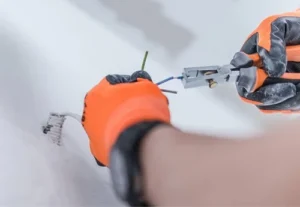 a close up of an electrician wearing orange safety gloves using electrical pliers to install new electrical wiring within a wall